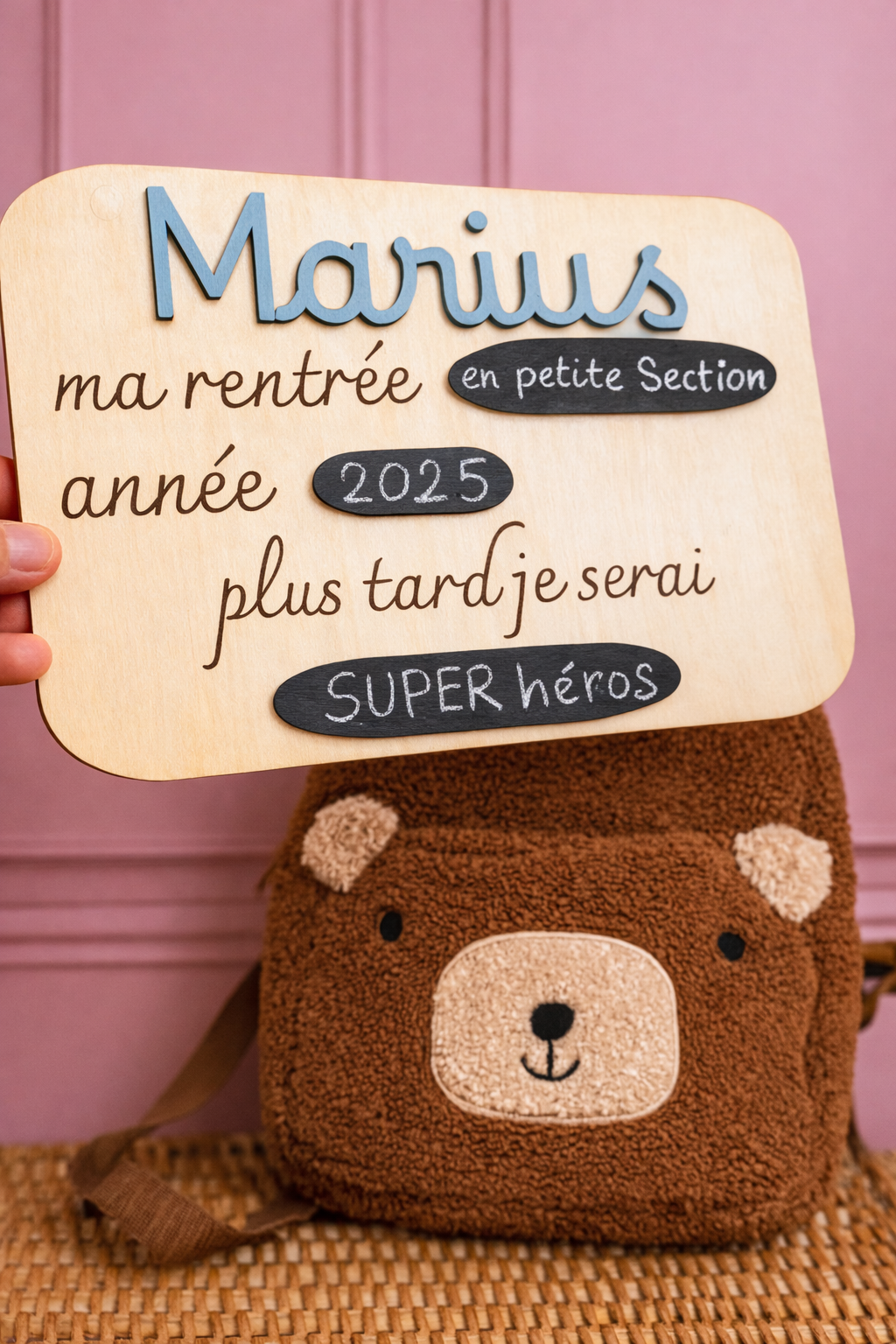 pancarte personnalisée pour photo rentrée scolaire avec prénom de l'"enfant peint et fixé sur la pancarte en bois (Marius en bleu) et avec 3 entrées gravées "ma rentrée" "année" "plus tard je serai" et 3 ardoises collées? la pancarte est tenue par la main au dessus d'un panier en osier surlequel est posé un sac à dos ourson, le mur est rose poudré