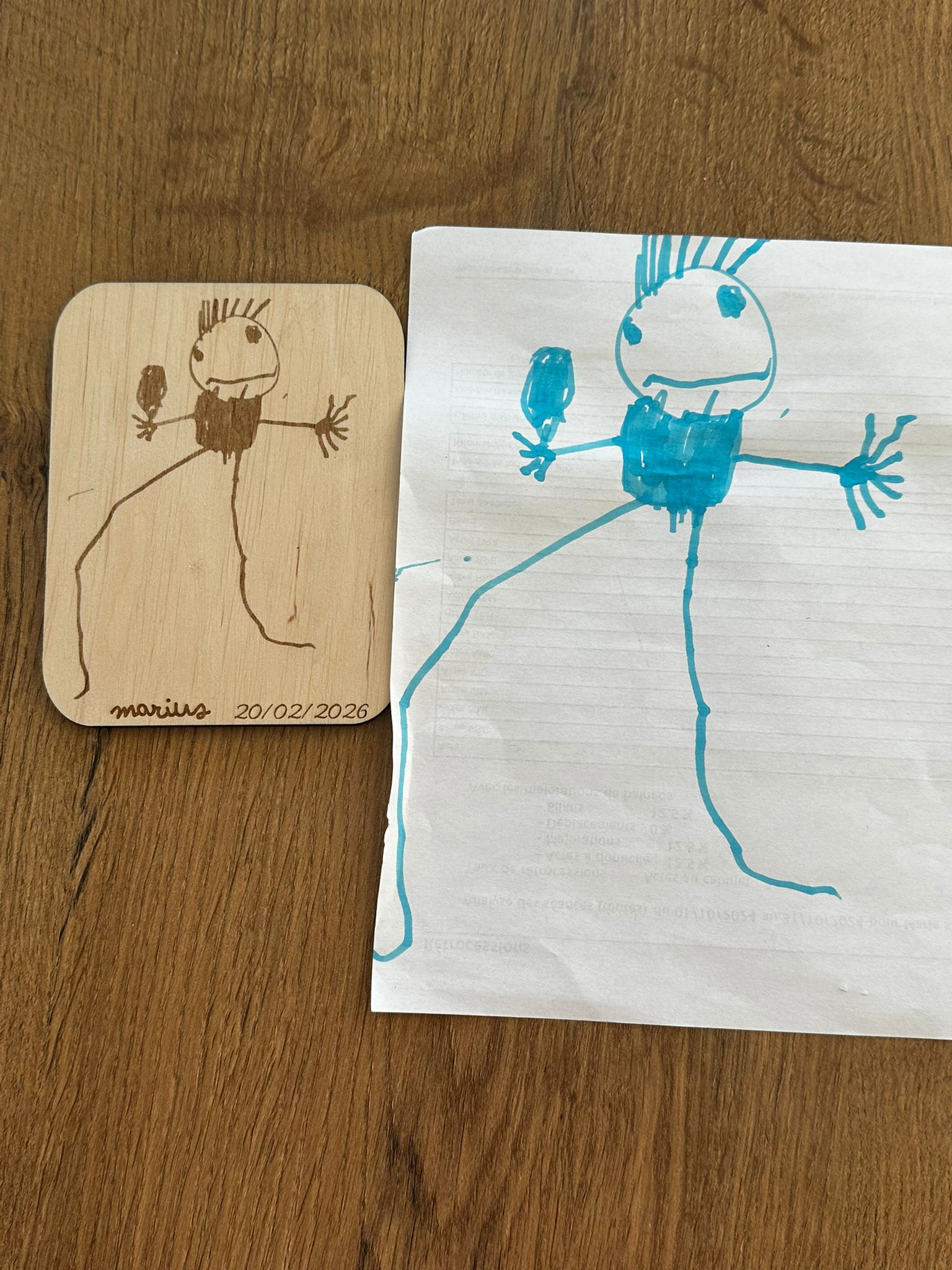 un dessin d'un bonhomme réalisé par un jeune enfant placé à côté du même dessin reproduit sur une carte en bois et signé "marius 20/02/2026"