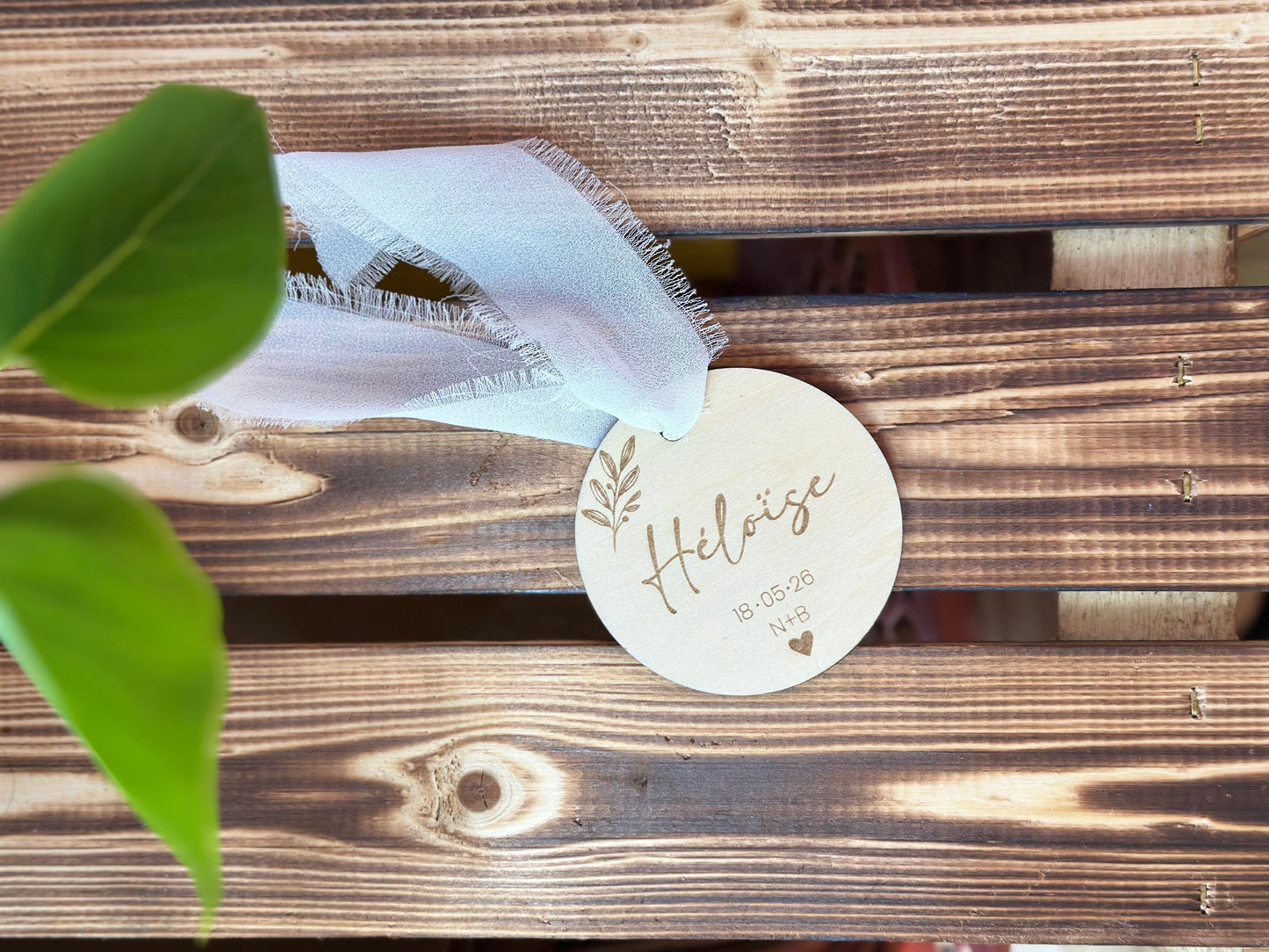 un marque place en bois personnalisé avec le prénom de l'invité gravé, la date du mariage, les initiales des mariés et des motifs végétaux. Posé sur une palette en bois et orné d'un ruban