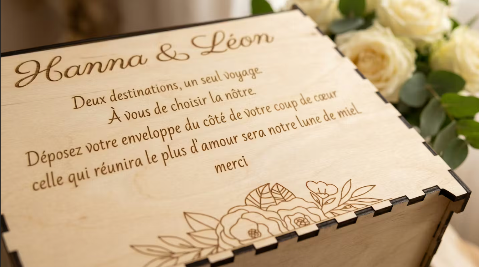 détail de l'urne en bois personnalisée pour le mariage d'Hanna & Léon avec un message personnalisé gravé dessus "deux destinations, un seul voyage. a vous de choisir la nôtre. déposez votre enveloppe du côté de votre coup de coeur, celle qui réunira le plus d'amour sera notre lune de miel. merci", urne posée prêt d'un bouquet de roses blanches