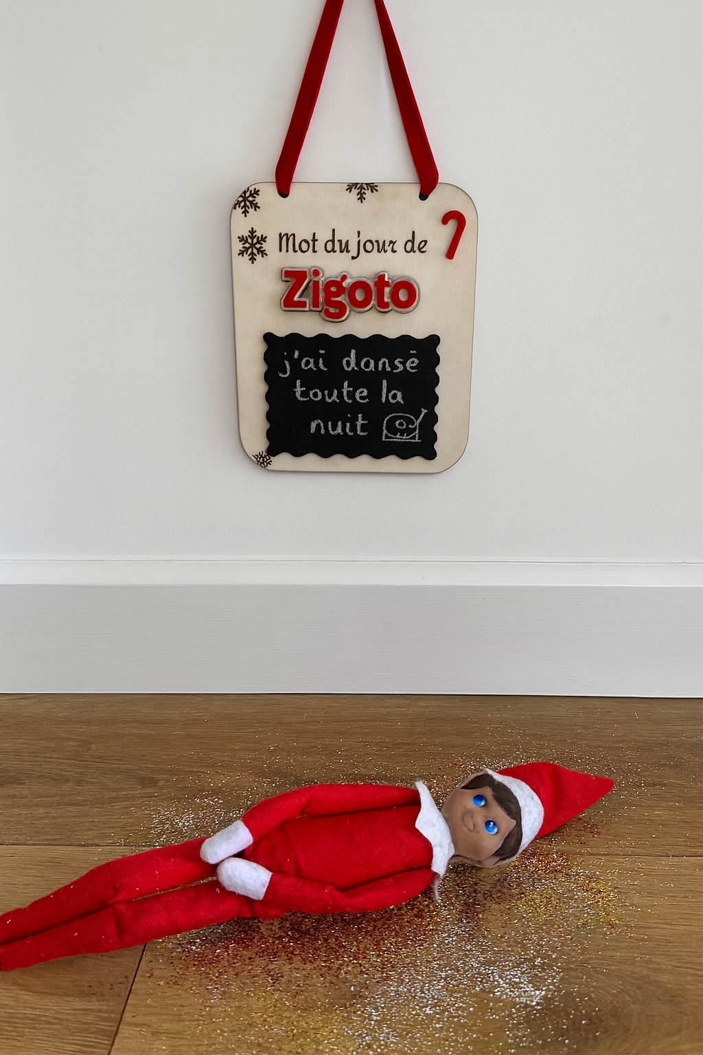 une pancarte en bois avec une ardoise pour que le lutin farceur puisse écrire un message chaque matin : ici : "j'ai dansé toute la nuit" avec un tourne disque dessiné. Suspenduau mur.  Prénom du lutin en plexi rouge personnalisé, ici "zygoto". l'elfe est allongé par terre entouré de paillettes