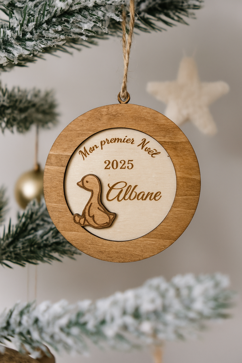 suspension ronde en bois en 3 dimensions : gravé "mon premier noël" puis "2025" et enfin le prénom de l'enfant "Albane" et détail d'un petit caneton vernis. La boule de noël personnalisée est accroché dans un sapin enneigé