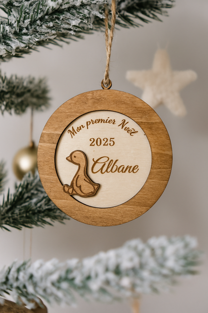 suspension ronde en bois en 3 dimensions : gravé "mon premier noël" puis "2025" et enfin le prénom de l'enfant "Albane" et détail d'un petit caneton vernis. La boule de noël personnalisée est accroché dans un sapin enneigé