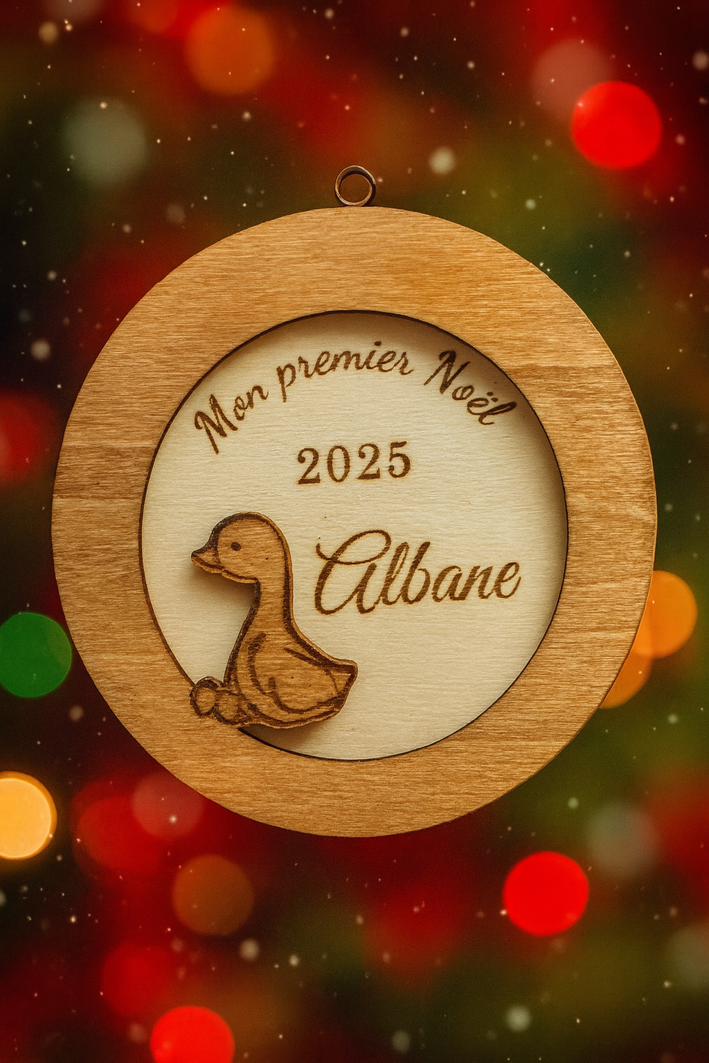 suspension ronde en bois en 3 dimensions : gravé "mon premier noël" puis "2025" et enfin le prénom de l'enfant "Albane" et détail d'un petit caneton vernis. La boule de noël personnalisée lévite, en fond des flocons de neige et des lumières de noël
