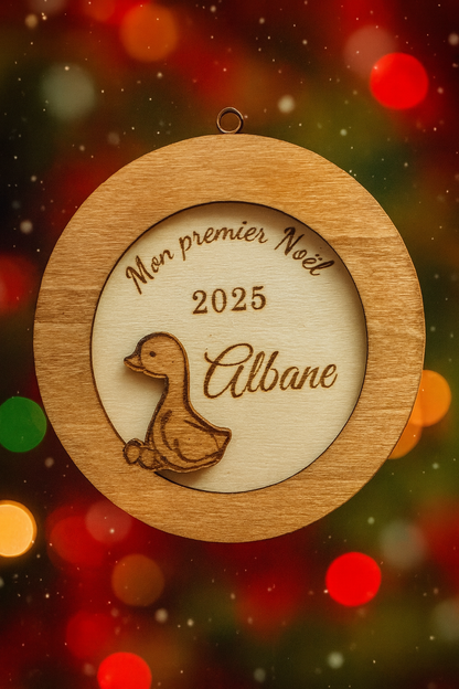 suspension ronde en bois en 3 dimensions : gravé "mon premier noël" puis "2025" et enfin le prénom de l'enfant "Albane" et détail d'un petit caneton vernis. La boule de noël personnalisée lévite, en fond des flocons de neige et des lumières de noël
