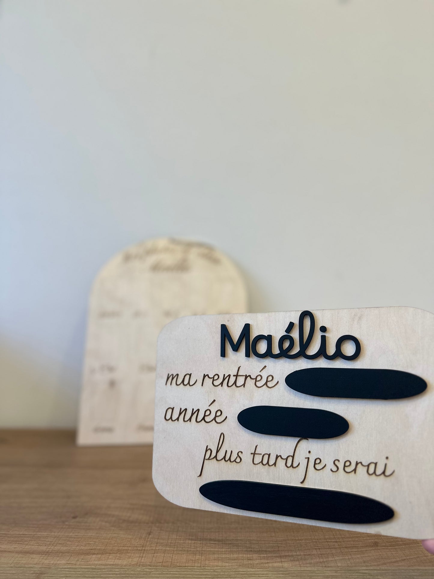 2 panneaux en bois pour le rituel de la rentrée scolaire : la pancarte ma rentrée avec l'année, la classe et le métier de rêve à remplir à la craie sur les ardoises ainsi que le prénom en relief peint en noir (ou autre couleur) personnalisé, ici "Maélio" puis le panneau mes photos de classe personnalisé avec le prénom de l'enfant "Maélio" et les emplacements pour le photos de la toute petite section à la 3eme. Les panneaux en bois sont posés sur un meuble en bois contre un ur blanc 
