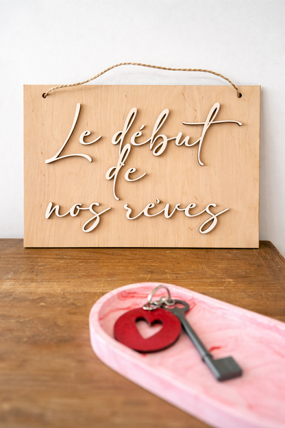 Pancarte en bois clair gravée “Le début de nos rêves” en lettres blanches 3D, posée sur un meuble en bois, avec un vide-poche rose et une clé flous au premier plan, lumière naturelle douce.