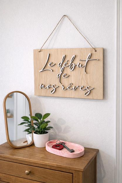 PPancarte en bois clair suspendue au-dessus d’un petit meuble d’entrée, accompagnée d’un miroir décoratif, d’une plante verte en pot blanc et d’un vide-poche rose, lumière naturelle élégante.