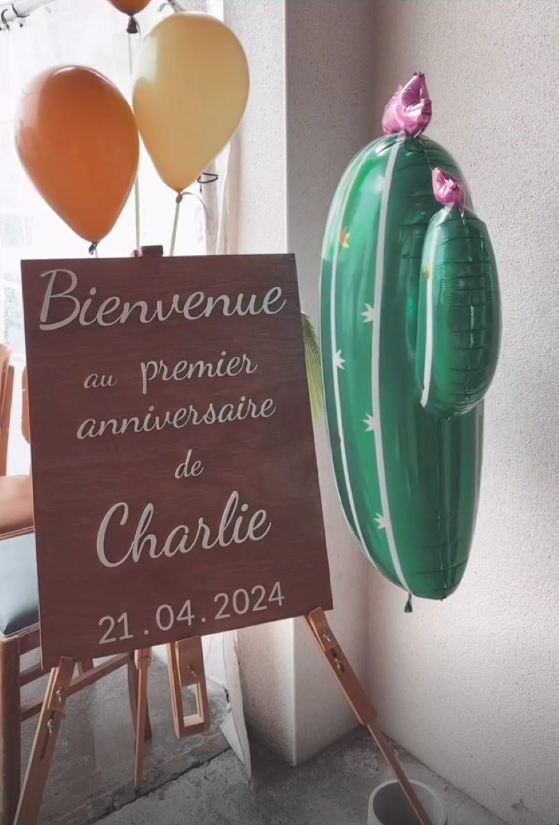 pancarte mariage ou autre évènement personnalisée en bois, "bienvenue au premier anniversaire de Charlie" peint et fixé sur le panneau en bois qui est posé sur un tretaux de tableau. autour des ballons (marrons, jaunes et un en forme de cactus"