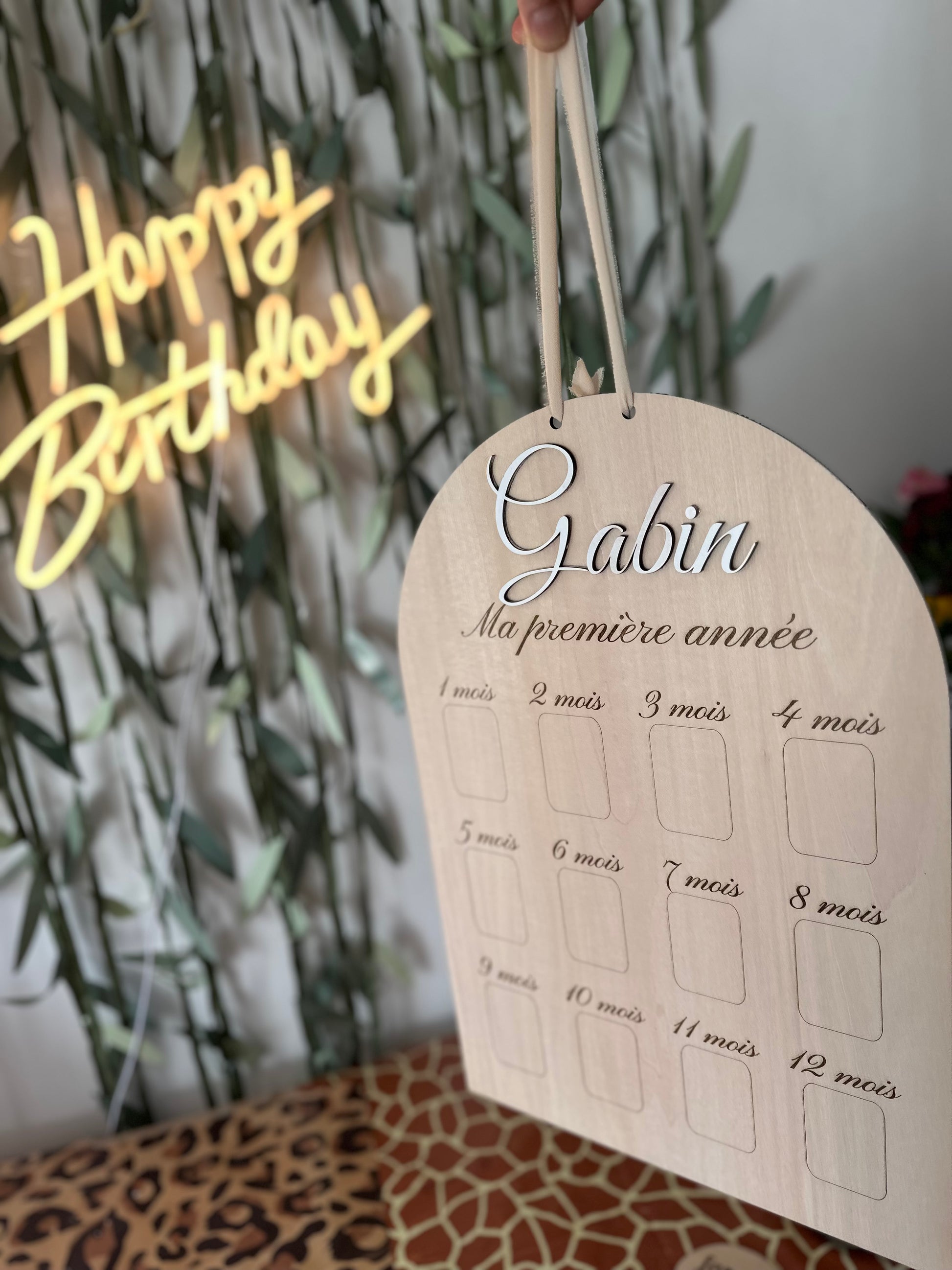 panneau en bois personnalisé "ma première année' avec le prénom de bébé "Gabin" et 12 emplacements pour y coller les photos de sa première année, en arrière plan un néon "happy birthday", une nappe léopard et girafe, et des lianes