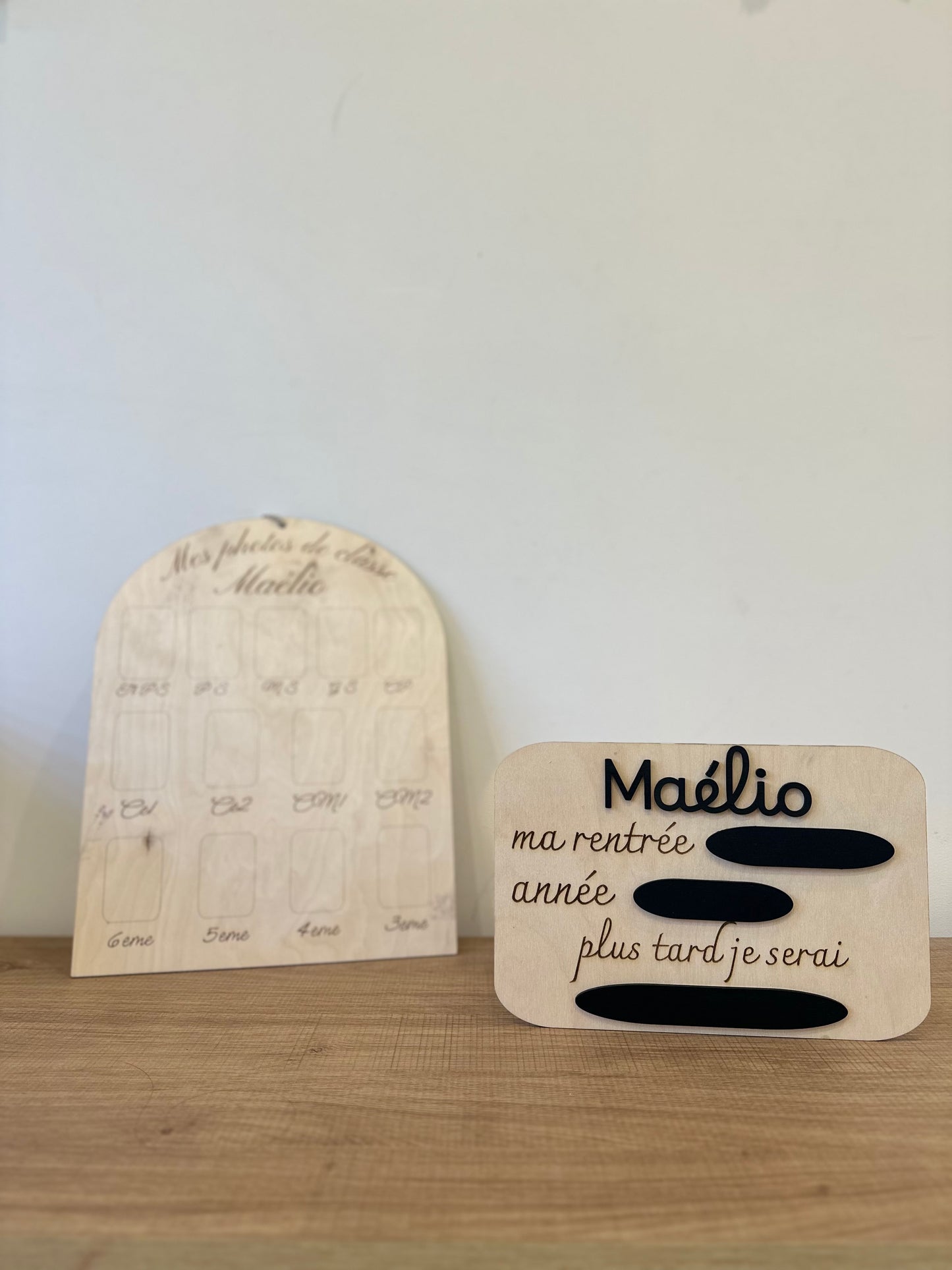 2 panneaux en bois pour le rituel de la rentrée scolaire : la pancarte ma rentrée avec l'année, la classe et le métier de rêve à remplir à la craie sur les ardoises ainsi que le prénom en relief peint en noir (ou autre couleur) personnalisé, ici "Maélio" puis le panneau mes photos de classe personnalisé avec le prénom de l'enfant "Maélio" et les emplacements pour le photos de la toute petite section à la 3eme. Les panneaux en bois sont posés sur un meuble en bois contre un ur blanc 