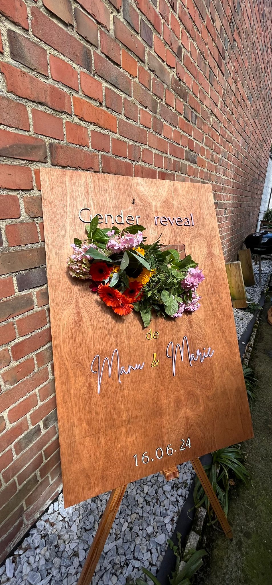 pancarte bienvenue évènement personnalisé/ le panneau en bois couleur chêne est posé en extérieur devant un beau mur de briques rouges et il est décoré de belles fleurs des champs et tournesols dans une encoche, il y a une inscription personnalisée "gender reveal" et "de Manu & Marie" ainsi que la date "16.06.24"