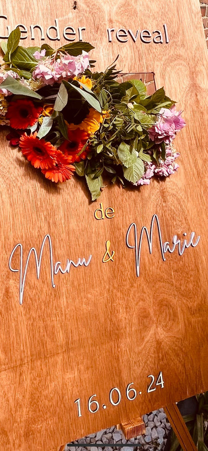 détail de la pancarte bienvenue personnalisée. L'écriteau en bois couleur chêne a une encoche pour laisser passer des fleurs des champs et tournesols et il y a une inscription personnalisée "gender reveal" et "de Manu & Marie" ainsi que la date "16.06.24"