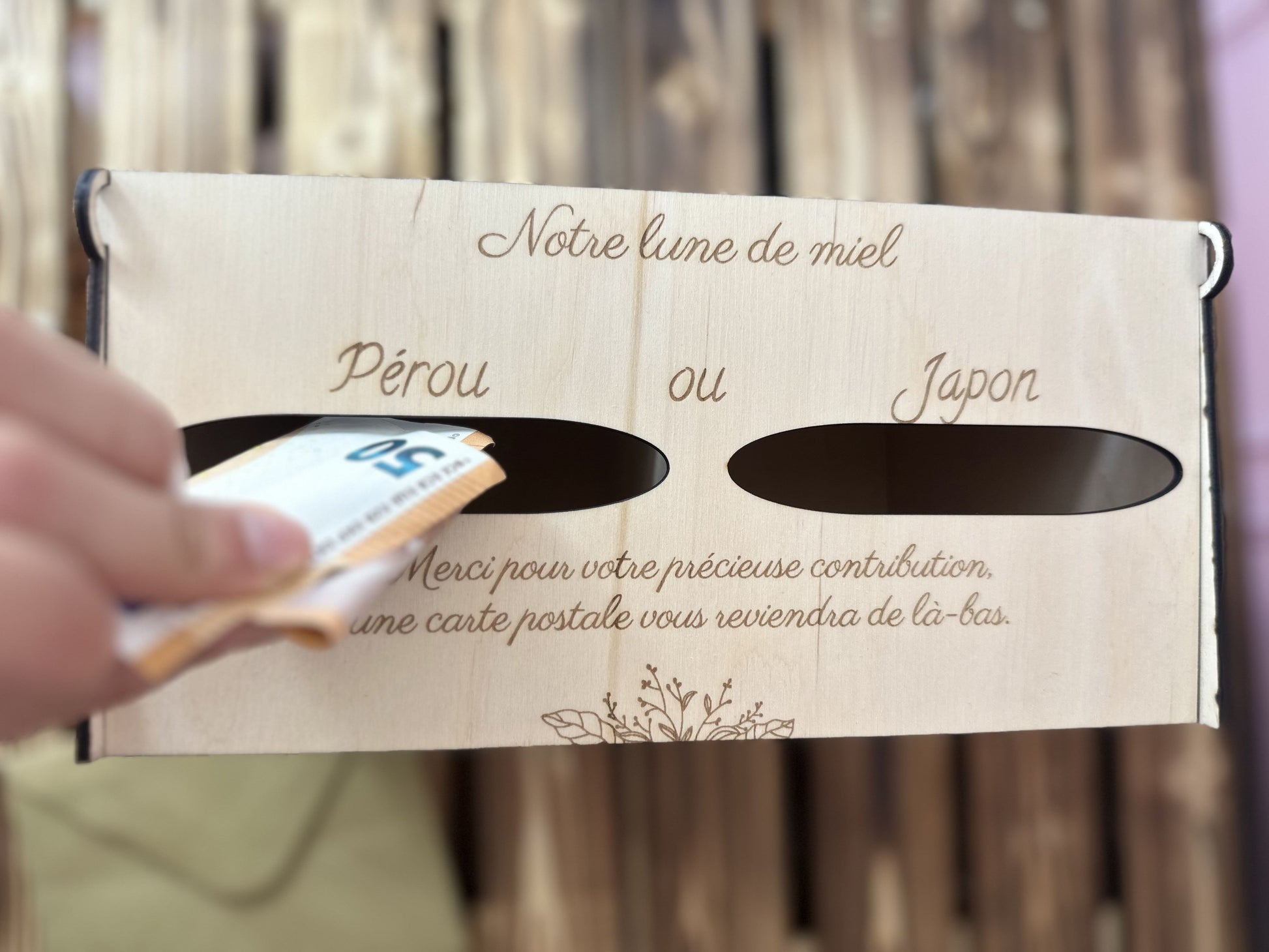 une urne en bois personnalisée pour pacs ou mariage avec un message gravé "notre lune de miel" 2 entrées pour voter "Pérou ou Japon" et un autre message gravé en dessous "merci pour votre contribution, une carte postale vous reviendra de là-bas" et une main glisse  billets de 50 euros dans l'entrée "Pérou", l'urne est vue du ciel, et posée sur une palette en bois