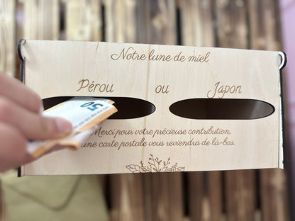 une urne en bois personnalisée pour pacs ou mariage avec un message gravé "notre lune de miel" 2 entrées pour voter "Pérou ou Japon" et un autre message gravé en dessous "merci pour votre contribution, une carte postale vous reviendra de là-bas" et une main glisse  billets de 50 euros dans l'entrée "Pérou", l'urne est vue du ciel, et posée sur une palette en bois