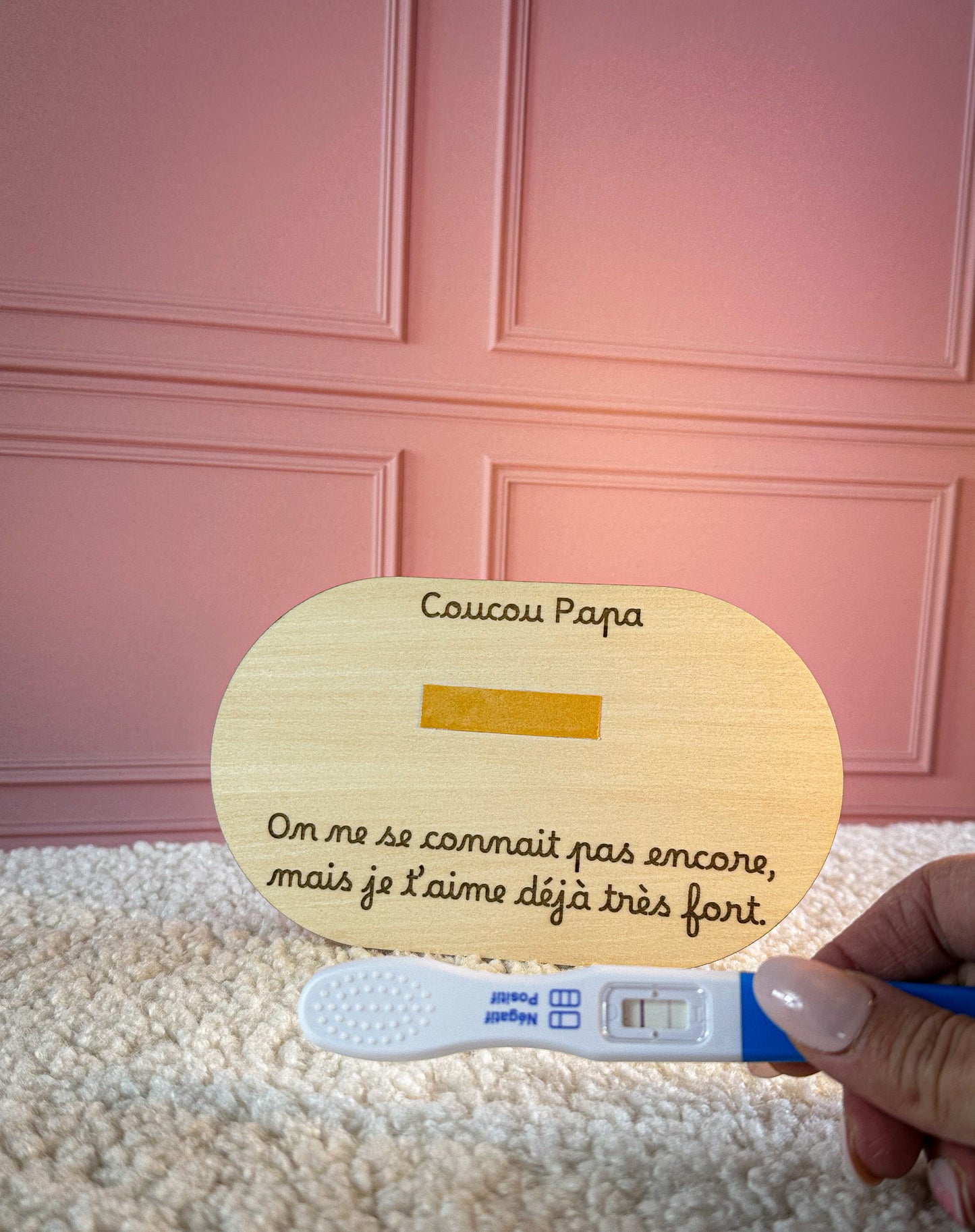 plaque annonce grossesse en bois avec emplacement pour test de grossesse. un main de femme tient un test de grossesse positif devant la carte en bois, la carte est posée sur un tapis blanc, en arrière plan un mur rose poudré
