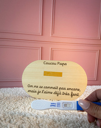 plaque annonce grossesse en bois avec emplacement pour test de grossesse. un main de femme tient un test de grossesse positif devant la carte en bois, la carte est posée sur un tapis blanc, en arrière plan un mur rose poudré