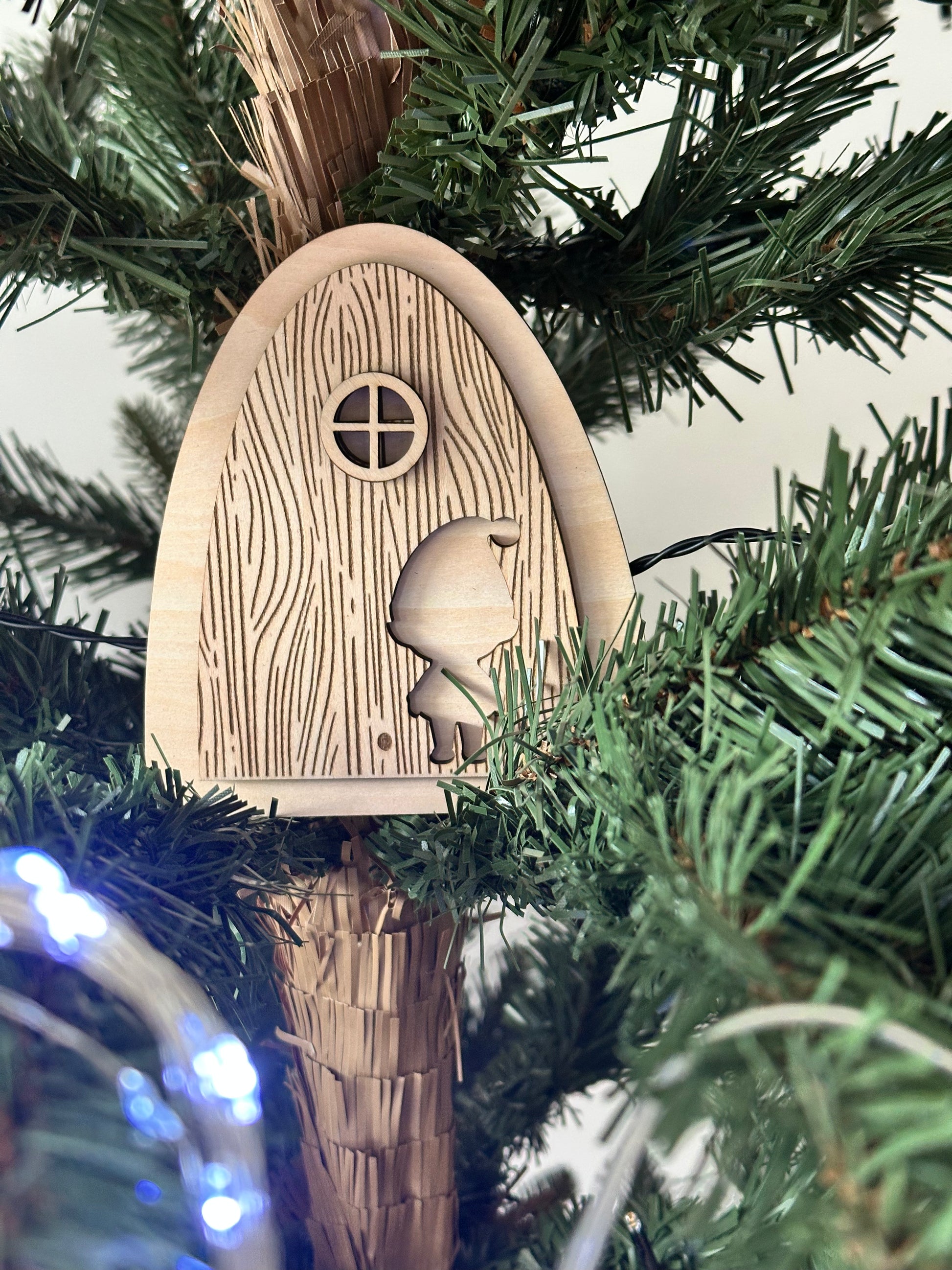 Porte de lutin farceur en bois suspendue dans un sapin de Noël, avec silhouette de lutin gravée et décorations lumineuses.