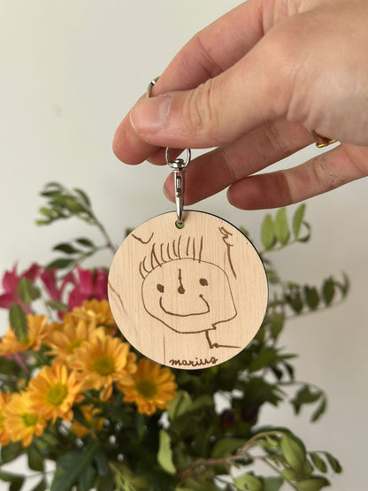une main adulte tient un porte clef sur lequel est reproduit un bonhomme dessiné par un enfant et signé par le prénom "marius" d'une écriture enfantine. en arrière plan un bouquet de fleurs jaunes et roses