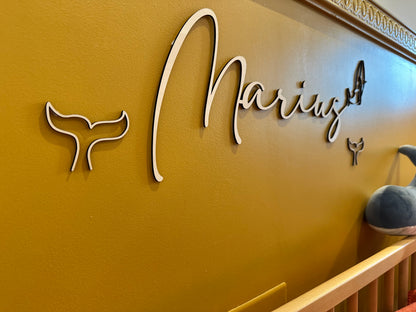 détails du prénom découpé au laser "Marius" se terminant par une baleine et entouré de queues de baleines. posé sur un mur jaune, au-dessus d'un lit d'enfant, avec une peluche baleine