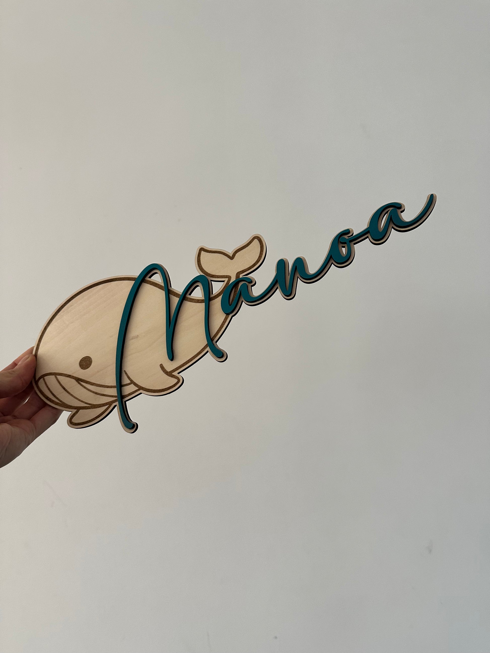 pancarte en bois en forme de baleine avec un prénom en bois peint en bleu fixé dessus. tenue par une main sur un fond mur blanc