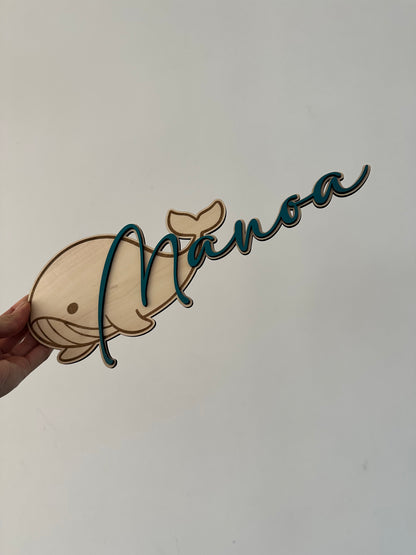 pancarte en bois en forme de baleine avec un prénom en bois peint en bleu fixé dessus. tenue par une main sur un fond mur blanc