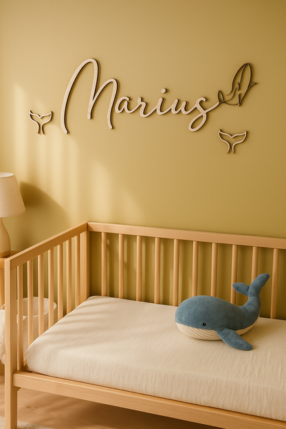 prénom en bois  "Marius" découpé au laser se terminant par une baleine et entouré de queues de baleine fixé sur un mur jaune moutarde au dessus d'un lit d'enfant sur lequel est posée une peluche baleine