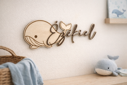 prénom en bois peint en marron "Ezékiel" et fixé sur une baleine en bois gravée pour une décoration de chambre sut le thème marin. La baleine est fixée sur un mur blanc, au dessus d'une étagère sur laquelle est posée une peluche baleine, un panier dont sort un linge bleu. Sur le côté  on aperçoit une illustration de baleine posée sur une petite étagère en bois