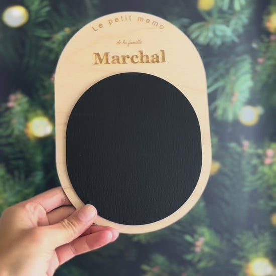 Vidéo du petit mémo personnalisé tenu dans une main : ardoise aimantée ovale en bois clair gravée au nom de famille, devant un sapin de Noël