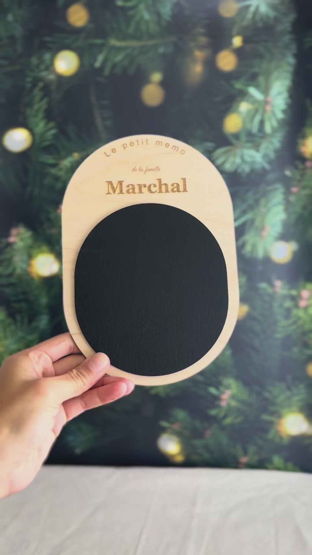 Vidéo du petit mémo personnalisé tenu dans une main : ardoise aimantée ovale en bois clair gravée au nom de famille, devant un sapin de Noël