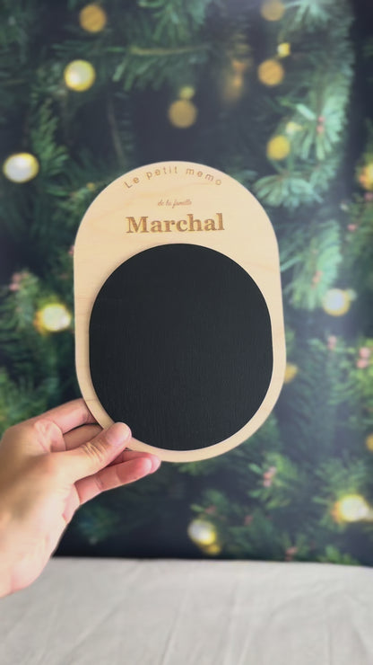 Vidéo du petit mémo personnalisé tenu dans une main : ardoise aimantée ovale en bois clair gravée au nom de famille, devant un sapin de Noël