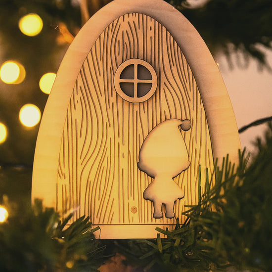 Porte du lutin farceur en bois naturel accrochée dans un sapin de Noël illuminé, entourée de guirlandes lumineuses dorées — décoration artisanale magique de Noël Chou Rose.