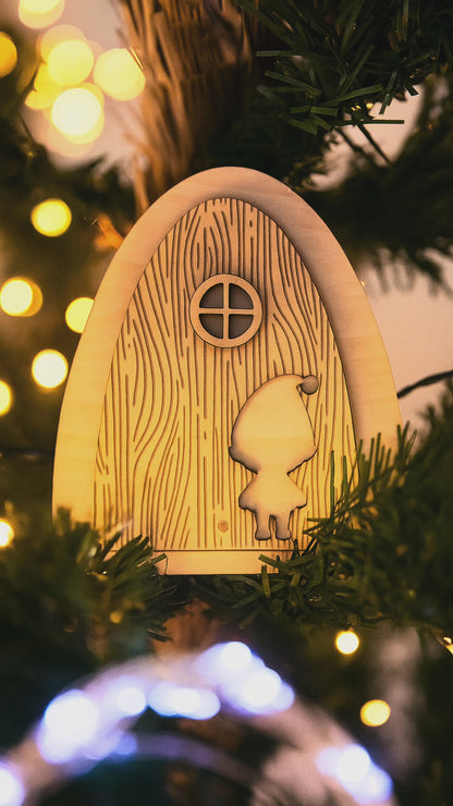 Porte du lutin farceur en bois naturel accrochée dans un sapin de Noël illuminé, entourée de guirlandes lumineuses dorées — décoration artisanale magique de Noël Chou Rose.