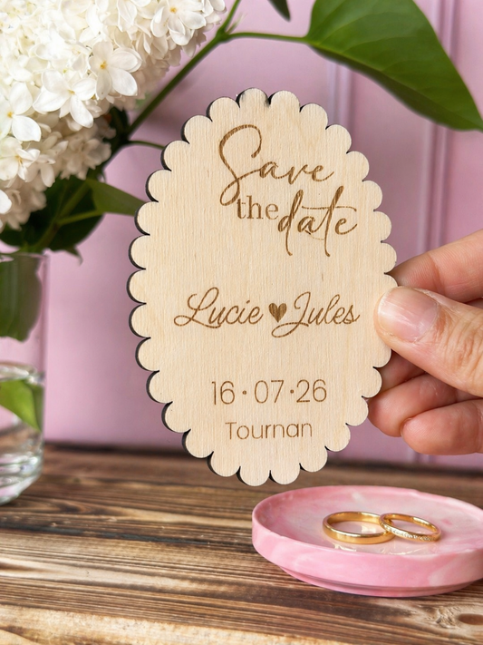 invitation mariage en bois personnalisée et gravée "save the date" avec les prénoms des futurs mariés "Lucie Jules" et la date ainsi que le lieu du mariage. Le médaillon est aimanté. on voit une paire d'alliance sur un plateau rose, un fond rose et une table en bois sur laquelle est posée du lilas