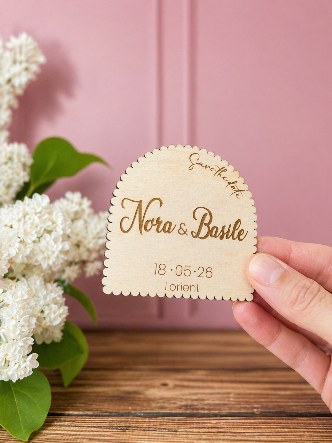 invitation mariage en bois en forme d'arche et personnalisée et gravée "save the date" avec les prénoms des futurs mariés "Nora & Basile" et la date ainsi que le lieu du mariage. Le médaillon est aimanté. Mur rose et moulé, table en bois massif et lilas blanc. L'invitation au mariage est tenue par une main féminine