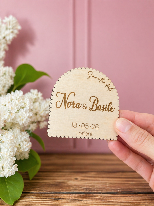 invitation mariage en bois en forme d'arche et personnalisée et gravée "save the date" avec les prénoms des futurs mariés "Nora & Basile" et la date ainsi que le lieu du mariage. Le médaillon est aimanté. Mur rose et moulé, table en bois massif et lilas blanc. L'invitation au mariage est tenue par une main féminine