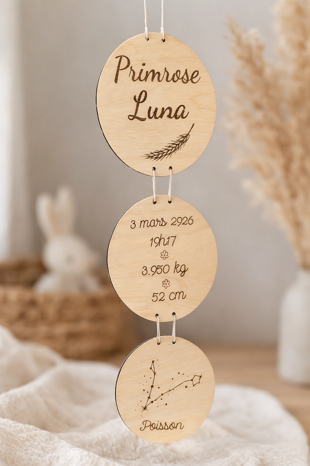 Mobile de naissance personnalisé en bois naturel avec trois disques gravés : le prénom « Primrose Luna », les informations de naissance et la constellation Poisson. Le mobile est suspendu dans une chambre bébé minimaliste aux tons beige et crème, entouré de fleurs séchées et de textiles en coton, dans une ambiance douce et artisanale.