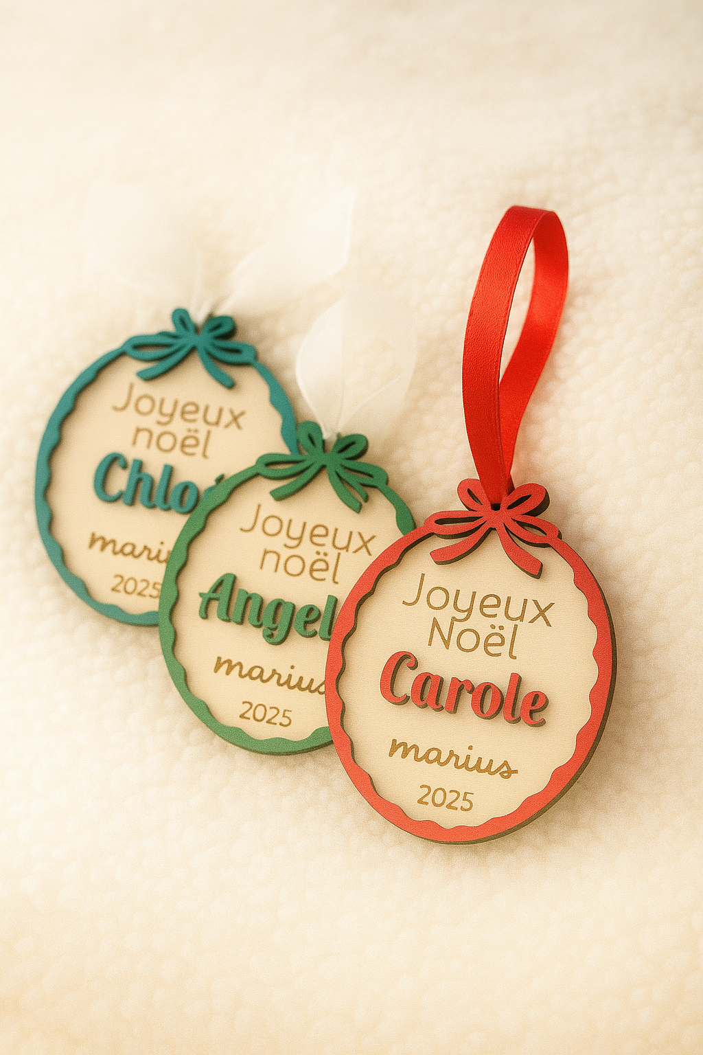 Trois suspensions de Noël personnalisées en bois gravé reposent sur un plaid en matière mouton. Chaque décoration, ornée d’un nœud coloré et d’un ruban, porte les inscriptions “Joyeux Noël” suivies des prénoms Carole, Angélique et Chloé, ainsi que “Marius 2025”. Les contours rouges, verts et bleus ajoutent une touche festive et artisanale, dans une ambiance douce et lumineuse signée Chou Rose.