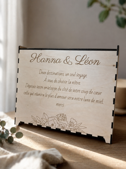 urne en bois personnalisée pour le mariage d'Hanna & Léon avec un message personnalisé gravé dessus "deux destinations, un seul voyage. a vous de choisir la nôtre. déposez votre enveloppe du côté de votre coup de coeur, celle qui réunira le plus d'amour sera notre lune de miel. merci", urne posée sur une table en bois et éclairée par la lumière du soleil à travers les rideaux
