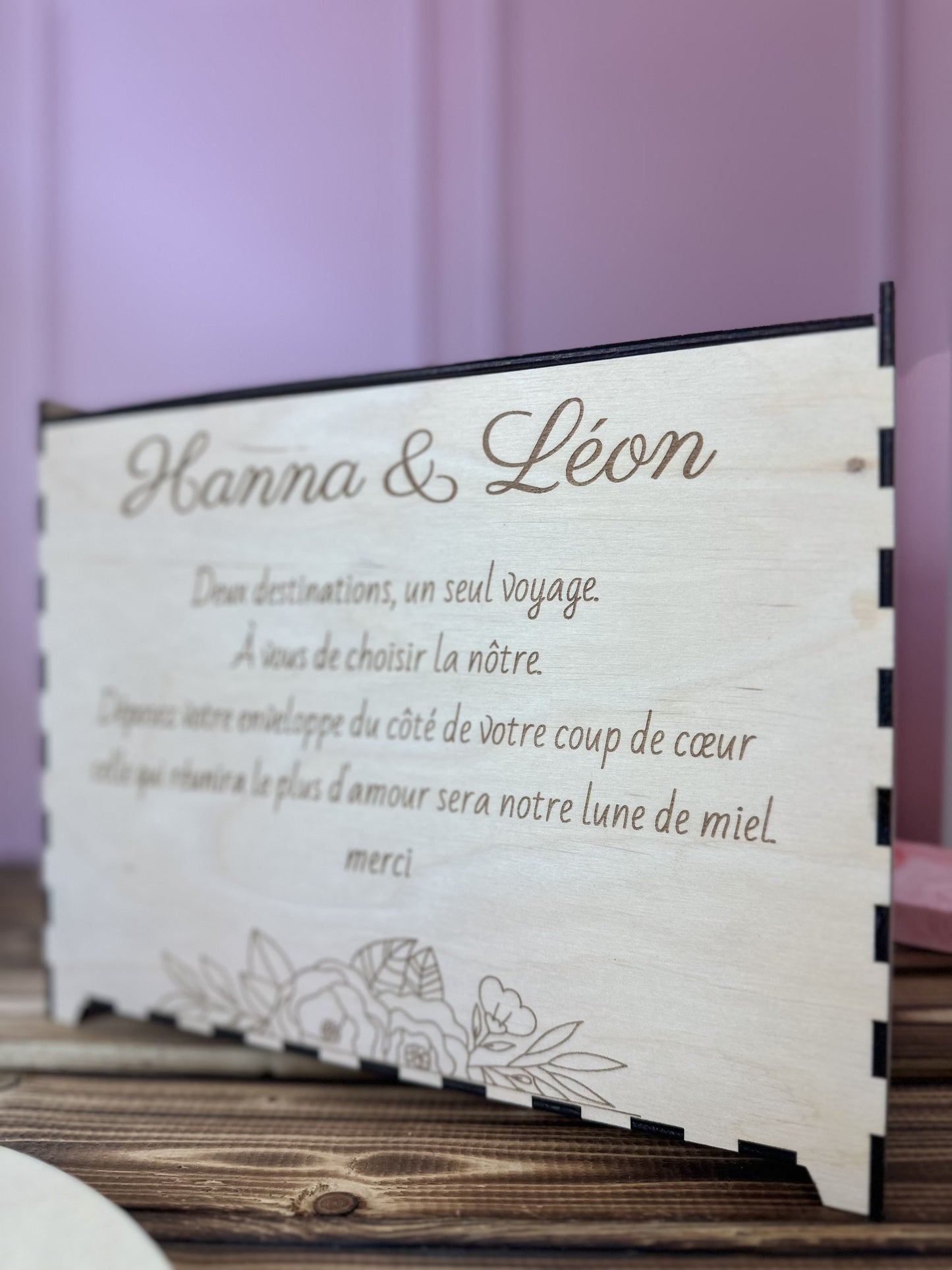 urne en bois personnalisée pour le mariage d'Hanna & Léon avec un message personnalisé gravé dessus "deux destinations, un seul voyage. a vous de choisir la nôtre. déposez votre enveloppe du côté de votre coup de coeur, celle qui réunira le plus d'amour sera notre lune de miel. merci", urne posée sur une table en bois, fond rose