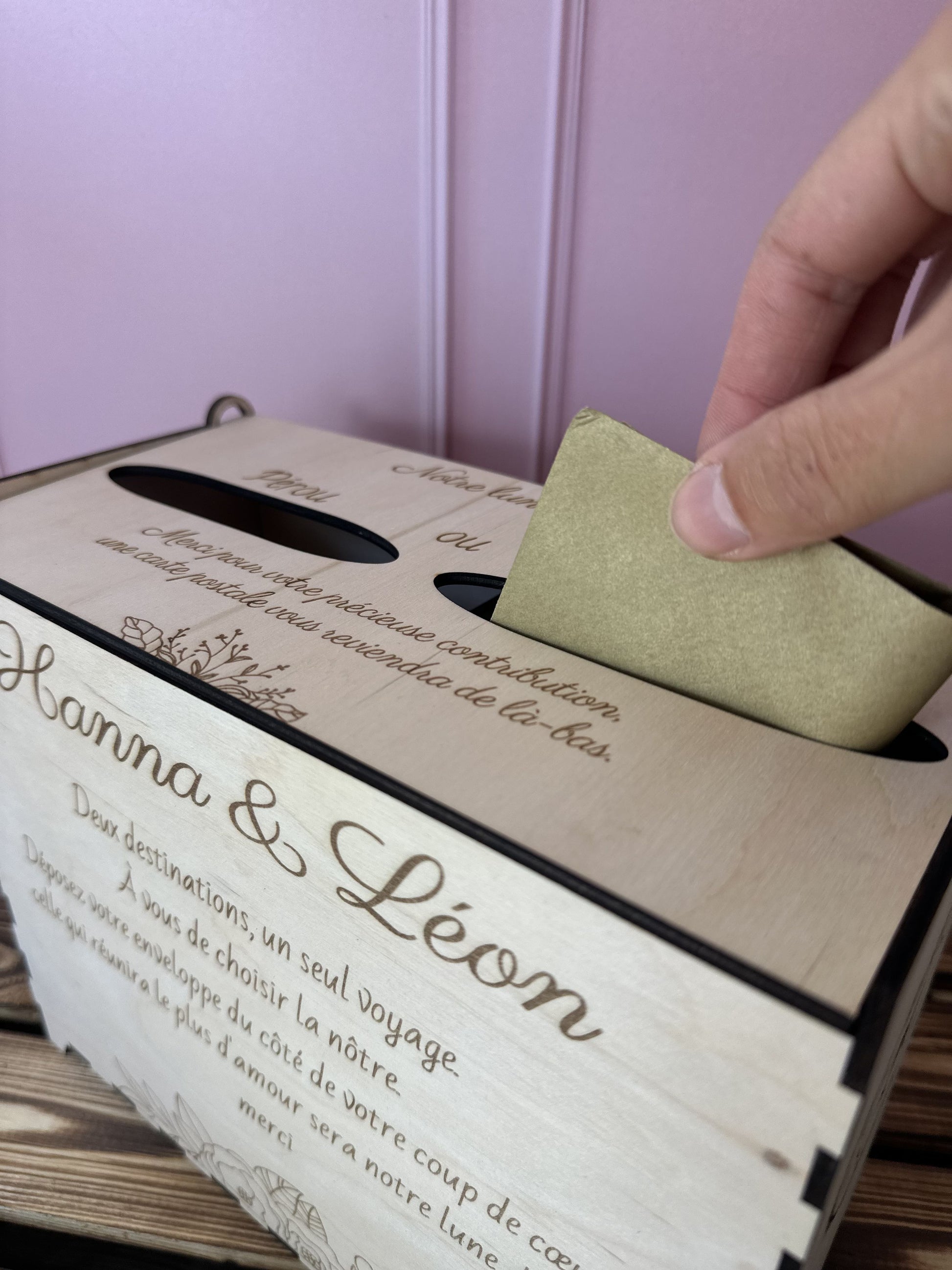 urne en bois personnalisée pour le mariage d'Hanna & Léon avec un message personnalisé gravé dessus "deux destinations, un seul voyage. a vous de choisir la nôtre. déposez votre enveloppe du côté de votre coup de coeur, celle qui réunira le plus d'amour sera notre lune de miel. merci", urne posée sur une table en bois, une main glisse une enveloppe dans l'une des 2 entrées à voter pour la destination