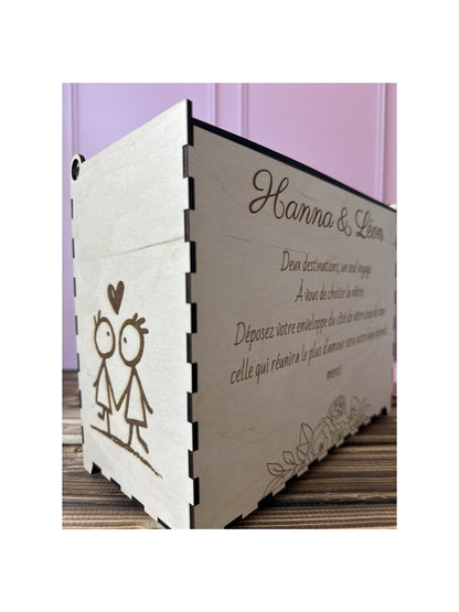 urne en bois personnalisée pour le mariage d'Hanna & Léon avec un message personnalisé gravé dessus "deux destinations, un seul voyage. a vous de choisir la nôtre. déposez votre enveloppe du côté de votre coup de coeur, celle qui réunira le plus d'amour sera notre lune de miel. merci", avec détail sur la tranche gauche de l'urne en bois : un dessin enfantin de 2 personnages
